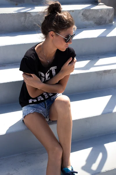 Young European teenage brunette curly model posing on stadium concrete stairs, wearing blue jeans mini shorts, sneakers, round sunglasses and black t-shirt, ready to party