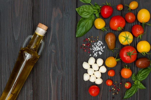 Cherry tomatoes of various color, mozzarella, basil leaves, spices and olive oil from above over dark wooden table. Italian caprese salad recipe ingredients. Top view, free text copy space