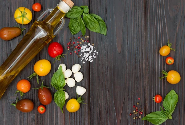 Cherry tomatoes of various color, mozzarella, basil leaves, spices and olive oil from above over dark wooden table. Italian caprese salad recipe ingredients. Top view, free text copy space