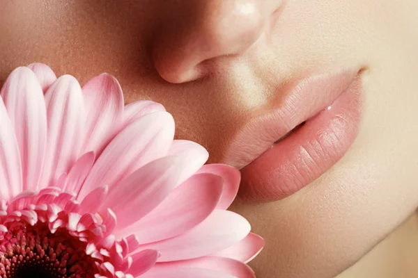 Lips with flower. Close-up beautiful female lips with bright lipgloss makeup. Perfect clean skin, light fresh lip make-up. Beautiful spa portrait with tender pink flower. Spa and cosmetics