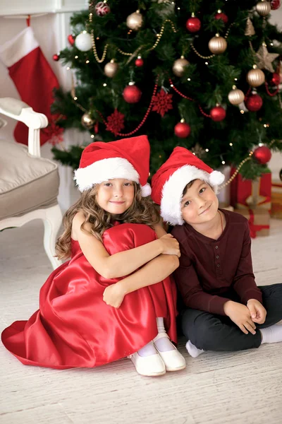 Children wearing Santa Claus sitting with gifts under the Christmas tree