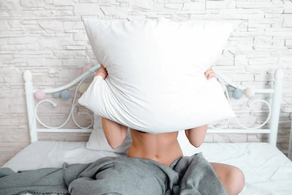 Naked woman hiding behind white pillow in bed