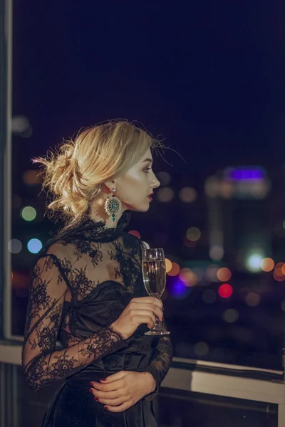 Pretty lady with glass of champagne looking through window to night city