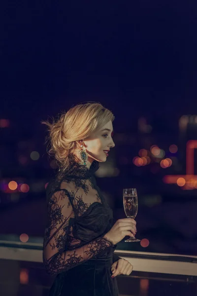 Pretty lady with glass of champagne looking through window to night city