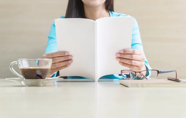 Asian woman reading a white book in her free time in the room with wooden desk and wall under window light , relaxation of asian woman concept by book coffee and eyeglasses