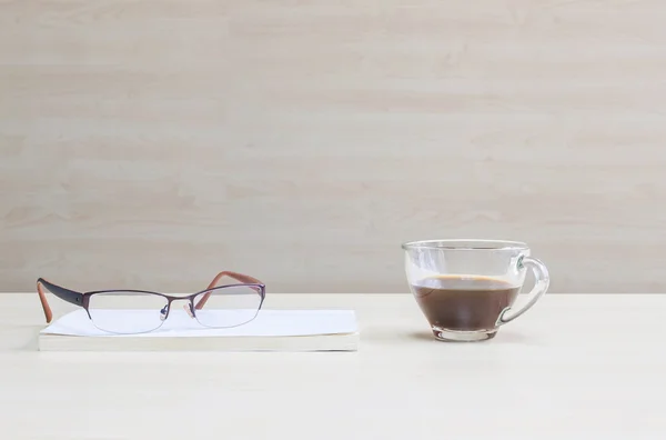 Closeup eyeglasses on white book with black coffee in transparent cup of coffee on blurred wooden desk and wall textured background in the meeting room under window