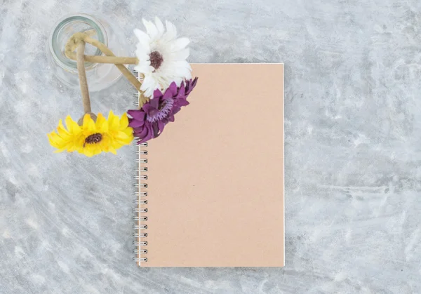 Closeup brown note book with blurred artificial colorful flower on transparent glass bottle on concrete table textured background in the garden in top view