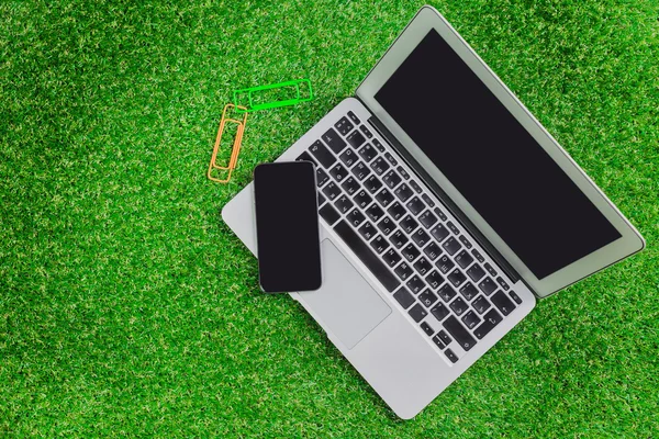 Blank notebook, phone, laptop and headphone on green grass