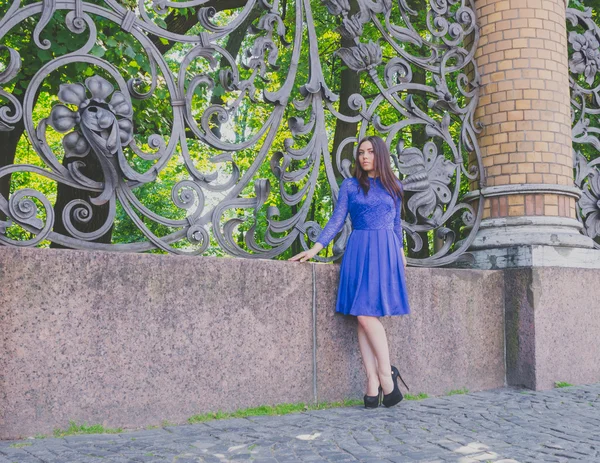 Beautiful girl in a blue dress stands near the old wrought-iron fence