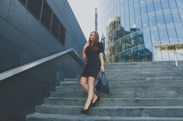 Beautiful girl in a black dress walks down the stairs holding a bag