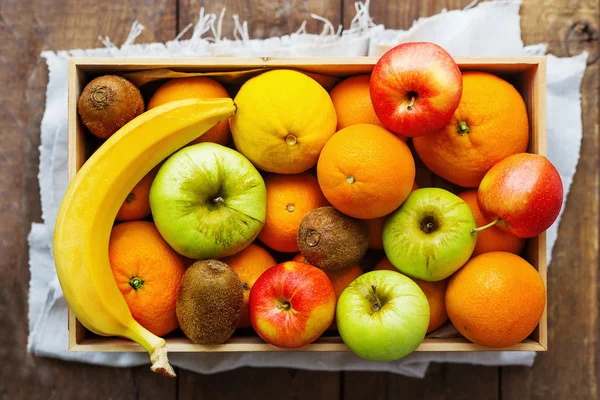 Box full of fresh fruits. Fruit harvest - apples, oranges, lemon, kiwi, banana. Rustic wooden table.