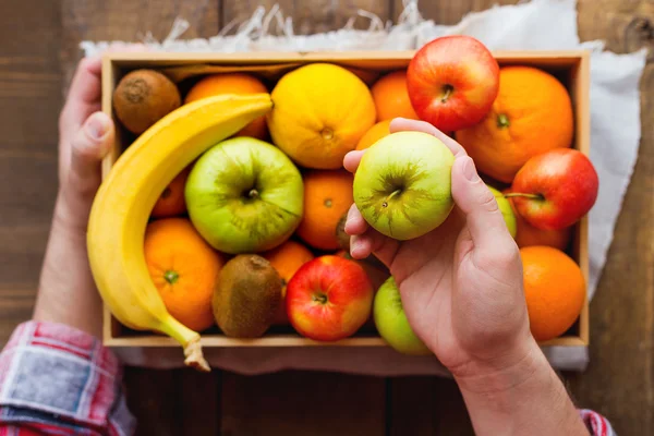 Man in tartan plaid shirt holds a box full of fresh fruits and a green apple. Fruit harvest - apples, oranges, lemon, kiwi, banana. Rustic wooden table.