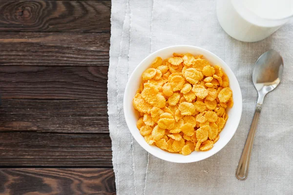 Tasty corn flakes in bowl with bottle of milk. Rustic wooden background with homespun napkin. Healthy crispy breakfast snack. Place for text. Top view, flat lay.