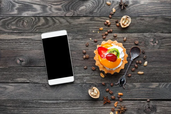 Wooden rustic background with fruit tart and spices - peanuts, anise stars, coffee beans, walnuts. Tasty dessert with strawberry, kiwi, orange, peach and whipped cream. Smartphone, good for mock up.