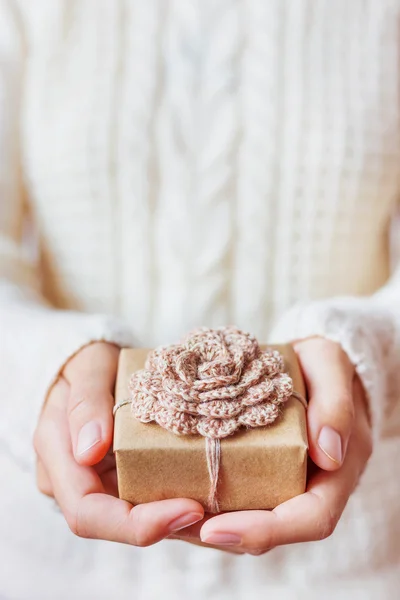 Woman in white knitted sweater holding a present. Gift is packed in craft paper with hand made crocheted flower. Example of DIY ways to pack Christmas and other presents.