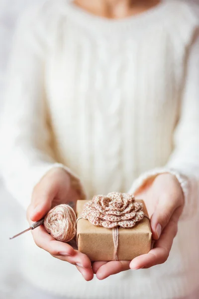 Woman in white knitted sweater holding a present and coil of thread with crochet hook. Gift is packed in craft paper with hand made crocheted flower. Example of DIY ways to pack Cristmas and other presents.