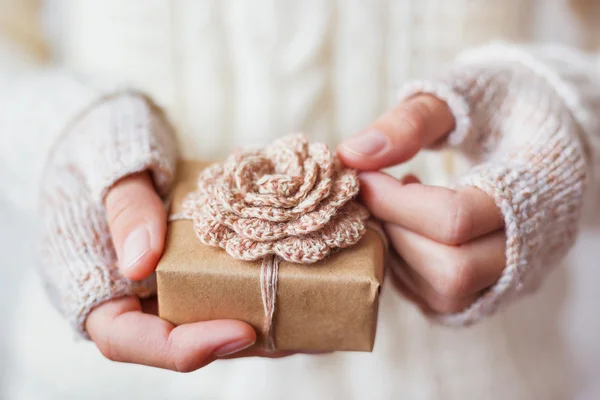 Woman in white knitted sweater and mitts holding a present. Gift is packed in craft paper with hand made crocheted flower. Example of DIY ways to pack Cristmas and other presents.