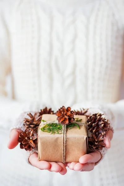 Woman in knitted sweater holding a present. Gift is packed in craft paper with pine cones and tied with rough rope. Hands full of pine cones. Example of DIY way to wrap a present.