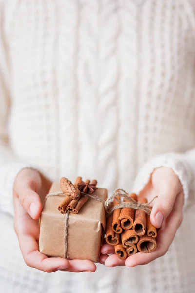 Woman in knitted sweater holding a present. Gift is packed in craft paper with vanilla pods, fir cone and star of anise.