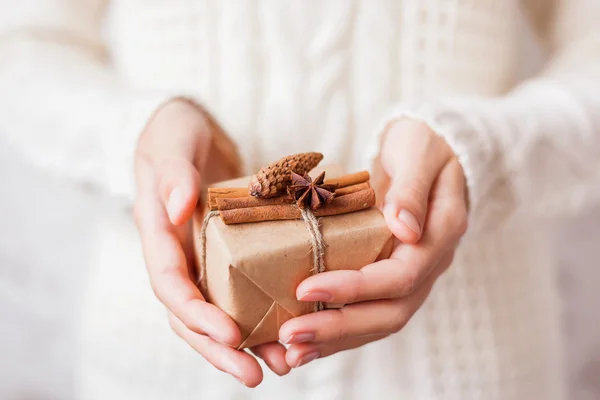 Woman in knitted sweater holding a present. Gift is packed in craft paper with vanilla pods, fir cone and star of anise.