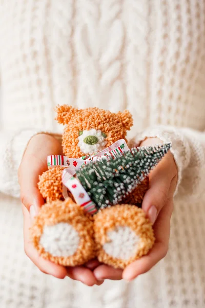 Woman in white knitted sweater holding toy bear with christmas ribbon and fir tree. Hand made crocheted little bear is a present for kid.