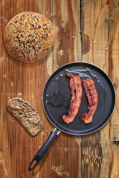 Fried Bacon Rashers in Teflon Frying Pan with Integral Bread Loaf and Slice on old Wooden Table