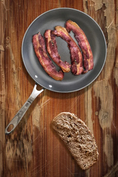 Fried Pork Bacon rashers in Teflon Frying Pan with slice of Bread on old Wooden Table