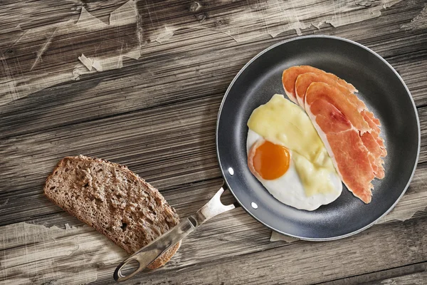 Prosciutto Rashers with Fried Egg and Cheese in Frying Pan with Bread Slice on Old Cracked Wooden Table