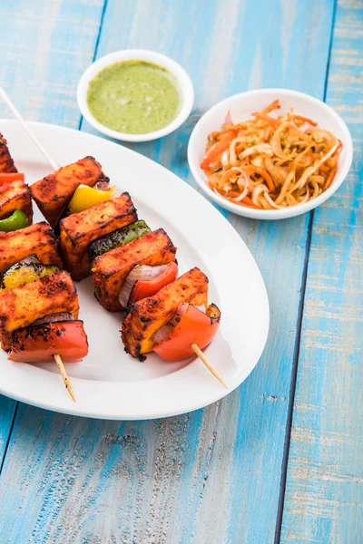 Paneer Tikka Kabab - Tandoori Indian cheese skewers, malai paneer tikka / malai paneer kabab, chilli paneer served in white plate with barbecue stick and colourful capsicum and onion, with green sauce