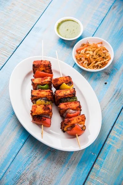 Paneer Tikka Kabab - Tandoori Indian cheese skewers, malai paneer tikka / malai paneer kabab, chilli paneer served in white plate with barbecue stick and colourful capsicum and onion, with green sauce