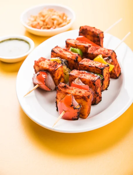 Paneer Tikka Kabab - Tandoori Indian cheese skewers, malai paneer tikka / malai paneer kabab, chilli paneer served in white plate with barbecue stick and colourful capsicum and onion, with green sauce