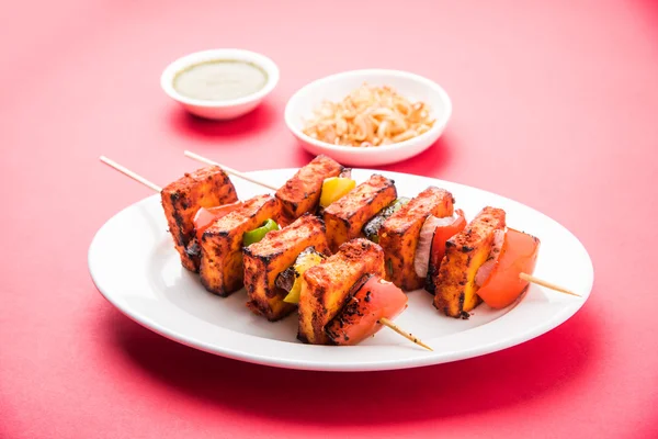 Paneer Tikka Kabab - Tandoori Indian cheese skewers, malai paneer tikka / malai paneer kabab, chilli paneer served in white plate with barbecue stick and colourful capsicum and onion, with green sauce