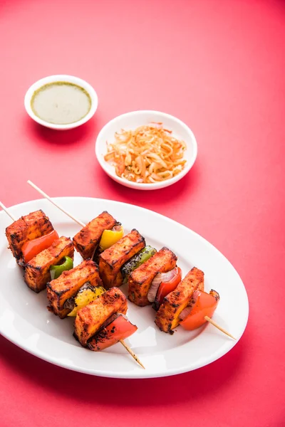 Paneer Tikka Kabab - Tandoori Indian cheese skewers, malai paneer tikka / malai paneer kabab, chilli paneer served in white plate with barbecue stick and colourful capsicum and onion, with green sauce