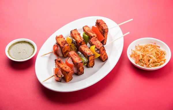 Paneer Tikka Kabab - Tandoori Indian cheese skewers, malai paneer tikka / malai paneer kabab, chilli paneer served in white plate with barbecue stick and colourful capsicum and onion, with green sauce