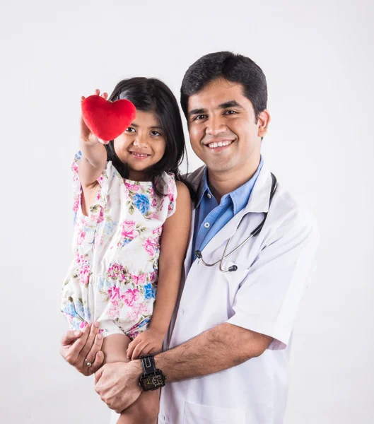 Handsome pediatric doctor holding a baby girl, male doctor with small girl, indian doctor, indian girl patient with red heart stuffed toy, heart care concept and doctor with girl patient, isolated