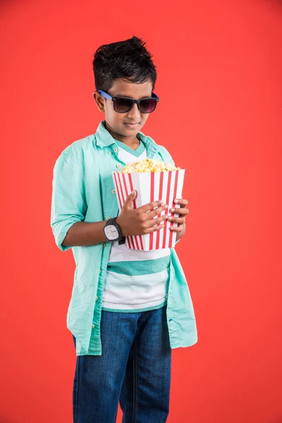 Joyful indian kid holding a big box of popcorn and looking at the camera isolated on yellow background, Portrait of adorable indian young boy eating popcorn, asian kid and popcorn