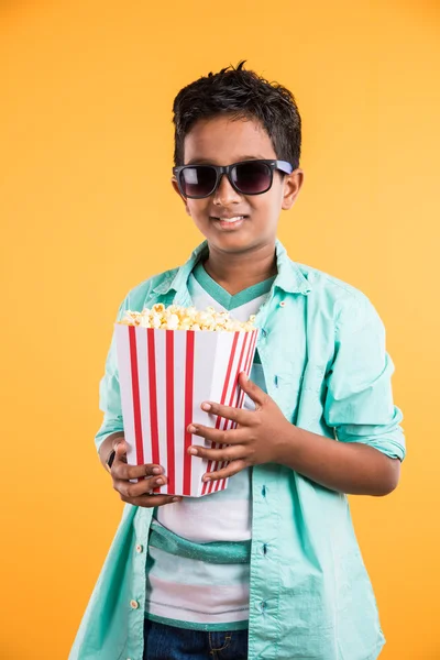 Joyful indian kid holding a big box of popcorn and looking at the camera isolated on yellow background, Portrait of adorable indian young boy eating popcorn, asian kid and popcorn