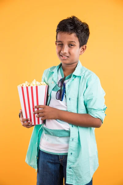 Joyful indian kid holding a big box of popcorn and looking at the camera isolated on yellow background, Portrait of adorable indian young boy eating popcorn, asian kid and popcorn