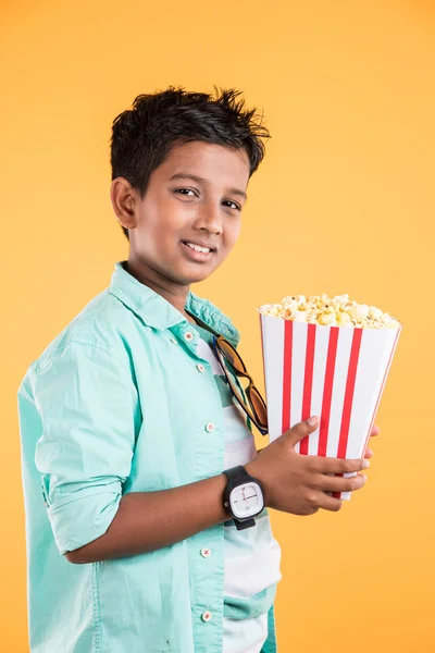 Joyful indian kid holding a big box of popcorn and looking at the camera isolated on yellow background, Portrait of adorable indian young boy eating popcorn, asian kid and popcorn