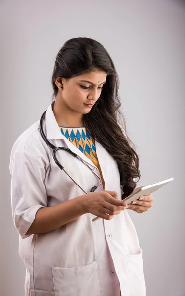 Beautiful Female Indian doctor looking at a digital tablet & wearing a white coat plus stethoscope