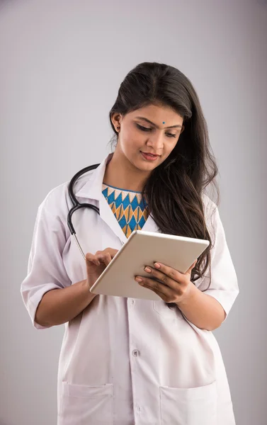 Beautiful Female Indian doctor looking at a digital tablet & wearing a white coat plus stethoscope