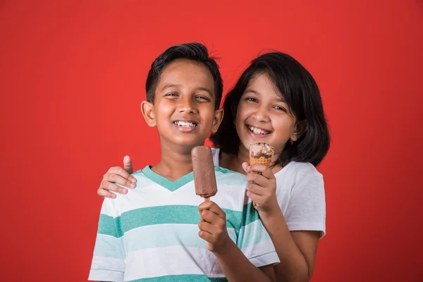 Two happy indian kids and ice cream, two asian kids enjoying ice cream or cone or chocolate candy, girl and boy eating ice cream, isolated on red background,