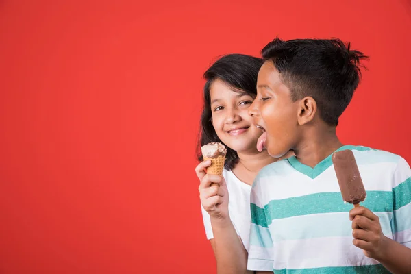 Two happy indian kids and ice cream, two asian kids enjoying ice cream or cone or chocolate candy, girl and boy eating ice cream, isolated on red background,