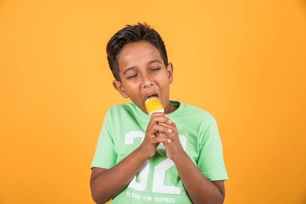 Little indian boy eating ice lolly or ice candy, indian boy eating mango ice cream, asian boy and orange ice candy, isolated on yellow background, 10 year boy and ice cream