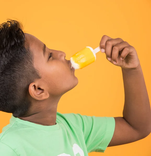 Little indian boy eating ice lolly or ice candy, indian boy eating mango ice cream, asian boy and orange ice candy, isolated on yellow background, 10 year boy and ice cream