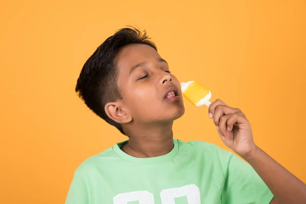 Little indian boy eating ice lolly or ice candy, indian boy eating mango ice cream, asian boy and orange ice candy, isolated on yellow background, 10 year boy and ice cream