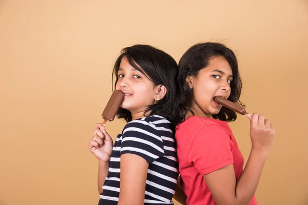 Two indian girls eating ice cream or ice candy, two asian girl and ice cream or ice candy, isolated on brown background, ten year old indian girls enjoying ice cream or ice candy or choko bar