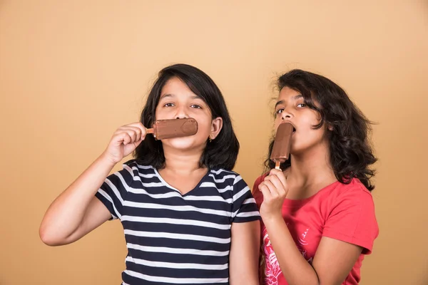 Two indian girls eating ice cream or ice candy, two asian girl and ice cream or ice candy, isolated on brown background, ten year old indian girls enjoying ice cream or ice candy or choko bar