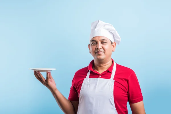 Portrait of an Indian Male cook in uniform holding an empty dish, presenting something. standing isolated over blue background, asian chef holding red plate