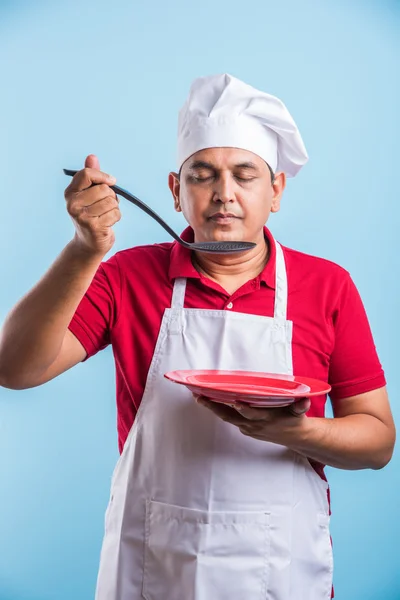 Portrait of an Indian male chef cook holding a red plate and ladle isolated on blue background, asian male chef tasting